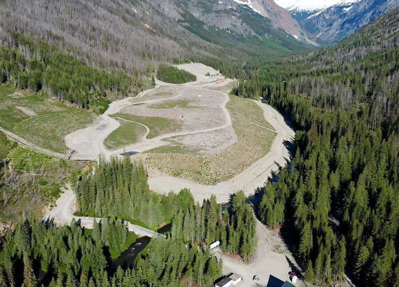 A mountain landscape showing a restored area with winding paths, surrounded by dense green forests and snow-capped peaks in the background. Showcasing the efforts of the restoration efforts in the closed mine.