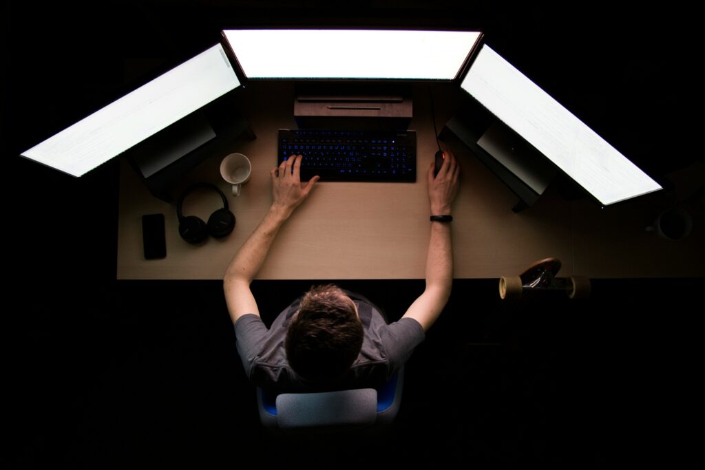 Overhead view of a person working at a desk with three bright computer monitors in a dark room, illustrating the relationship between technology, AI and the human mind.